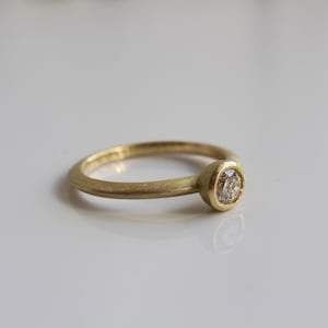 May include: A gold ring with a single, round diamond set in a bezel. The ring has a simple, elegant design, with a smooth, matte finish. The diamond is clear and sparkling, and the ring is set against a white background.