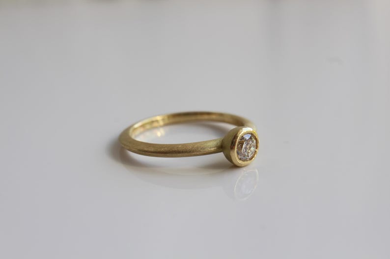 May include: A gold ring with a single, round diamond set in a bezel. The ring has a simple, elegant design, with a smooth, matte finish. The diamond is clear and sparkling, and the ring is set against a white background.