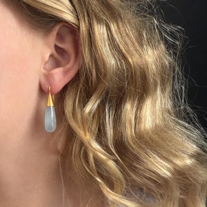 May include: Gold-tone earrings with a teardrop-shaped, pale blue gemstone. The earrings are hanging from a person's ear, with the gold hook visible. The background shows blonde hair and a white top.