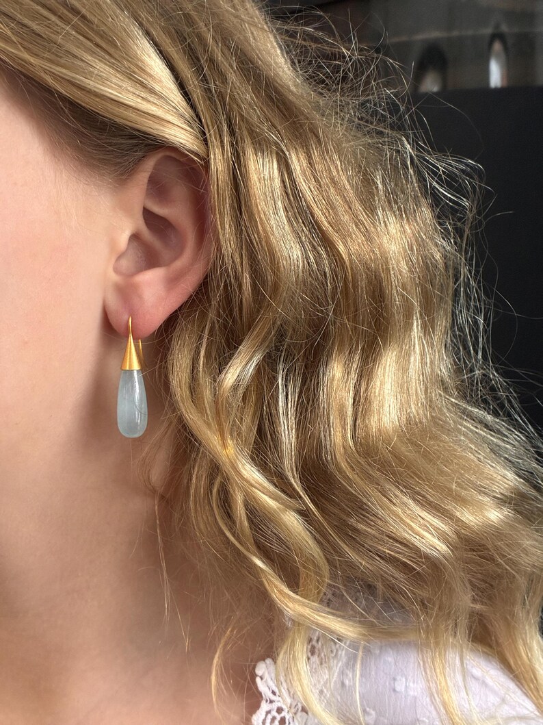 May include: Gold-tone earrings with a teardrop-shaped, pale blue gemstone. The earrings are hanging from a person's ear, with the gold hook visible. The background shows blonde hair and a white top.