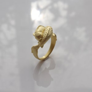 May include: A gold-colored ring featuring a koi fish design. The fish is sculpted in detail, with scales and fins visible. The ring's band curves around the fish, creating a unique and artistic piece of jewelry.