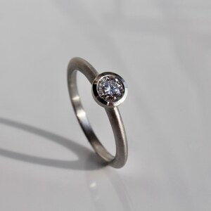 May include: A silver ring with a single, round, clear gemstone set in a bezel. The ring band is thin and smooth, and the gemstone is the focal point. The ring is displayed on a white surface, with a shadow.