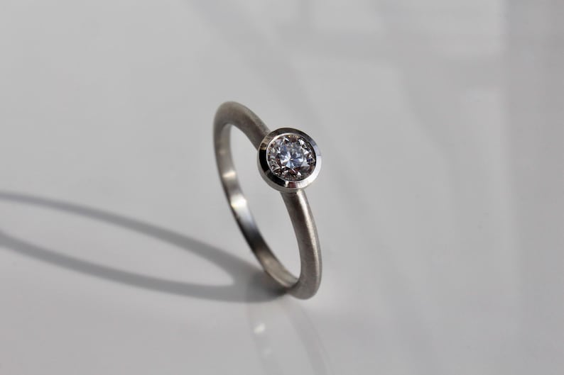 May include: A silver ring with a single, round, clear gemstone set in a bezel. The ring band is thin and smooth, and the gemstone is the focal point. The ring is displayed on a white surface, with a shadow.