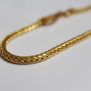 May include: A gold-toned chain bracelet featuring a dense, woven wheat-style link pattern. The metallic surface exhibits a polished finish with intricate, interlocking V-shaped segments that create a continuous, rounded, and flexible rope-like structure.