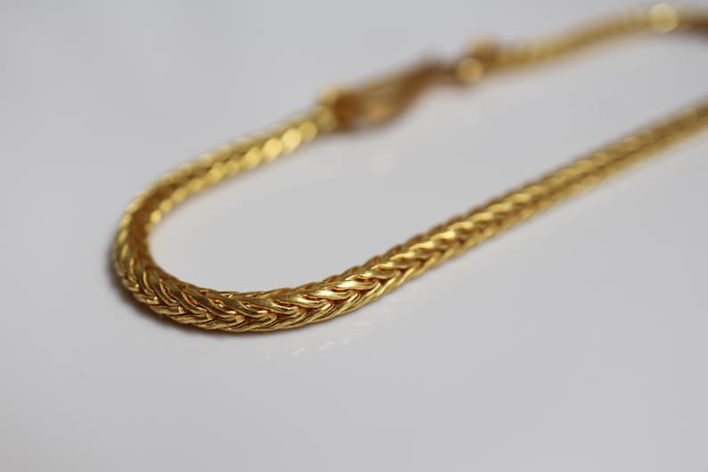 May include: A gold-toned chain bracelet featuring a dense, woven wheat-style link pattern. The metallic surface exhibits a polished finish with intricate, interlocking V-shaped segments that create a continuous, rounded, and flexible rope-like structure.