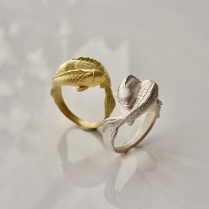 May include: Two rings with fish designs. One ring is gold-colored, and the other is silver-colored. The fish are sculpted with detailed scales and fins, and the rings have a smooth, polished finish. The rings are displayed on a white surface.