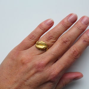May include: A gold-colored ring with a textured design, shaped like a stylized alligator or crocodile. The ring is worn on a finger, showcasing its intricate details and metallic sheen. The ring is a piece of jewelry.
