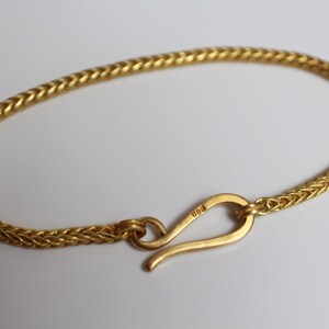 May include: A gold-colored bracelet featuring a woven wheat chain design. The bracelet is secured with a curved hook-style clasp that has the number 900 engraved on its surface. The metal has a consistent yellow tone throughout the entire piece.