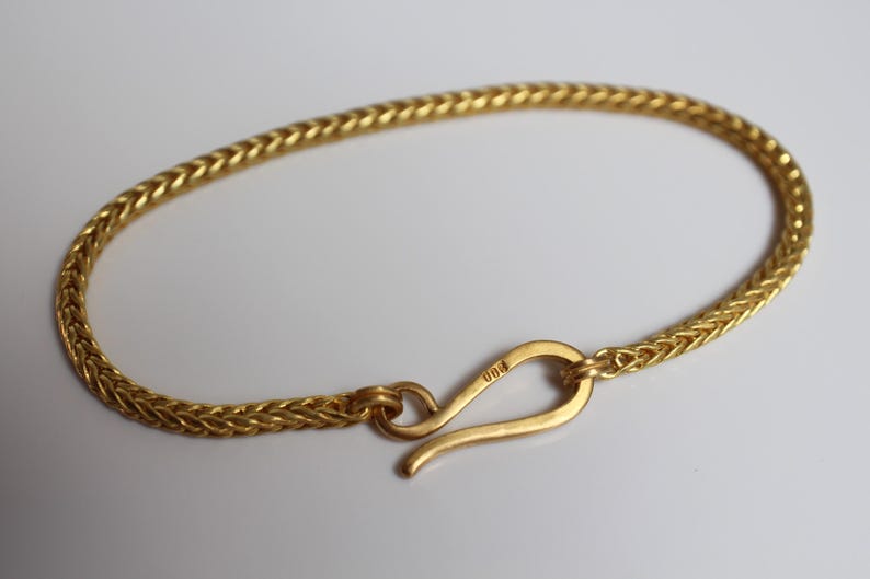 May include: A gold-colored bracelet featuring a woven wheat chain design. The bracelet is secured with a curved hook-style clasp that has the number 900 engraved on its surface. The metal has a consistent yellow tone throughout the entire piece.