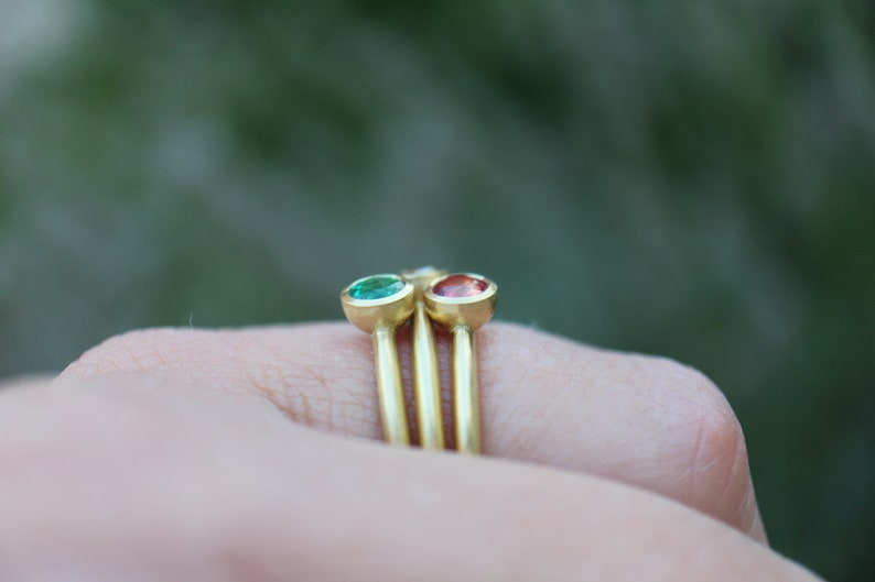May include: Three gold rings with bezel-set gemstones. One ring features a green gemstone, another a red gemstone, and the third a clear gemstone. The rings have thin, gold bands and are displayed against a blurred green background.