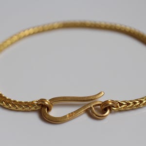 May include: A gold-colored bracelet featuring a woven chain design and a hook-style clasp. The clasp is stamped with the number 900. The bracelet is circular in shape and has a matte finish, showcasing a consistent metallic texture throughout the piece.