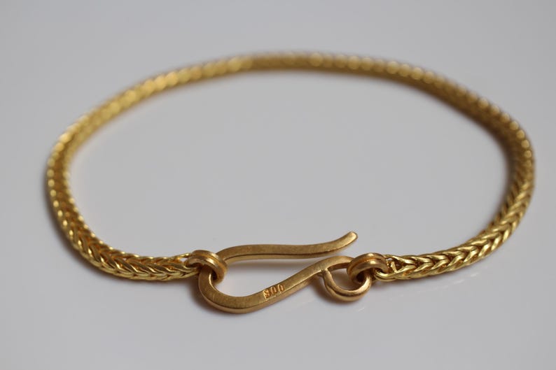 May include: A gold-colored bracelet featuring a woven chain design and a hook-style clasp. The clasp is stamped with the number 900. The bracelet is circular in shape and has a matte finish, showcasing a consistent metallic texture throughout the piece.
