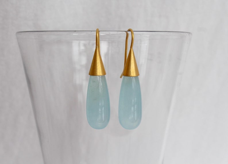 May include: A pair of elegant earrings with teardrop-shaped, pale blue gemstones. Each earring features a gold-colored conical cap and a curved hook. The earrings are displayed against a soft, neutral background, highlighting their delicate design.
