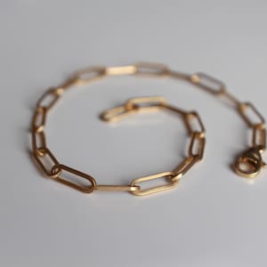 May include: A gold-toned chain bracelet featuring elongated, rectangular paperclip-style links. The bracelet is arranged in a circular shape and includes a secure lobster claw clasp mechanism at one end, showcasing a polished metallic finish.