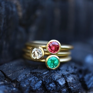 May include: Three gold rings with colorful gemstones. One ring features a clear diamond, another a red ruby, and the third a green emerald. The rings are stacked on a dark, textured surface.