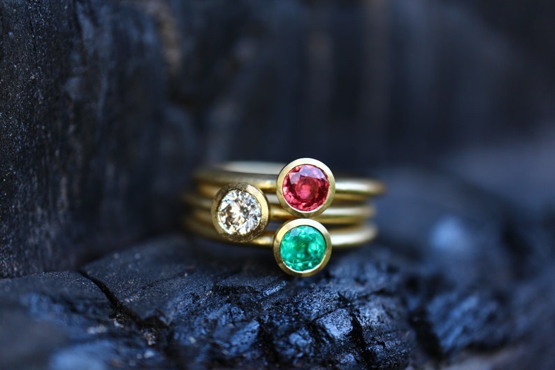 May include: Three gold rings with colorful gemstones. One ring features a clear diamond, another a red ruby, and the third a green emerald. The rings are stacked on a dark, textured surface.
