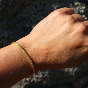 May include: A thin, gold-colored bracelet with a braided or wheat-chain texture. The bracelet is circular and rests on a wrist, displaying a metallic finish and a consistent, intricate woven pattern throughout the entire length of the jewelry piece.