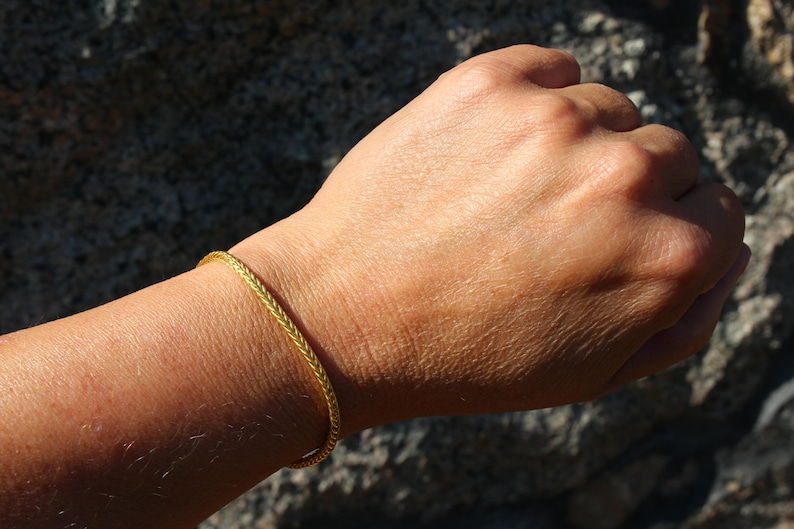 May include: A thin, gold-colored bracelet with a braided or wheat-chain texture. The bracelet is circular and rests on a wrist, displaying a metallic finish and a consistent, intricate woven pattern throughout the entire length of the jewelry piece.