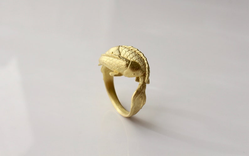 May include: A gold-colored ring featuring a koi fish design. The fish is sculpted with detailed scales and fins, wrapping around the band. The ring is set against a white background.