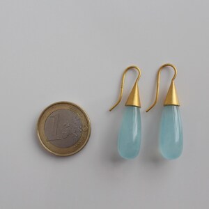 May include: A pair of gold-colored drop earrings with teardrop-shaped, light blue stones. The earrings are next to a one Euro coin. The earrings have a gold-colored conical cap and a curved hook.