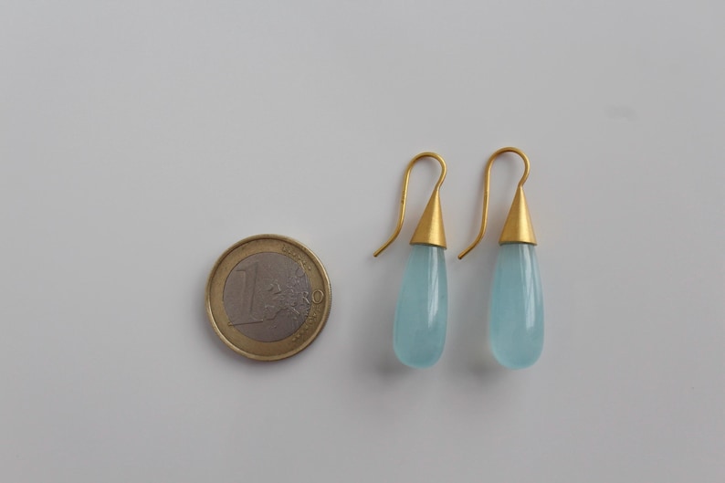 May include: A pair of gold-colored drop earrings with teardrop-shaped, light blue stones. The earrings are next to a one Euro coin. The earrings have a gold-colored conical cap and a curved hook.
