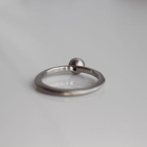 May include: A silver-toned metal ring featuring a single, small, spherical bead set at the top of the band. The ring has a smooth, polished finish and a simple, circular design that sits upright on a reflective, light-colored surface.