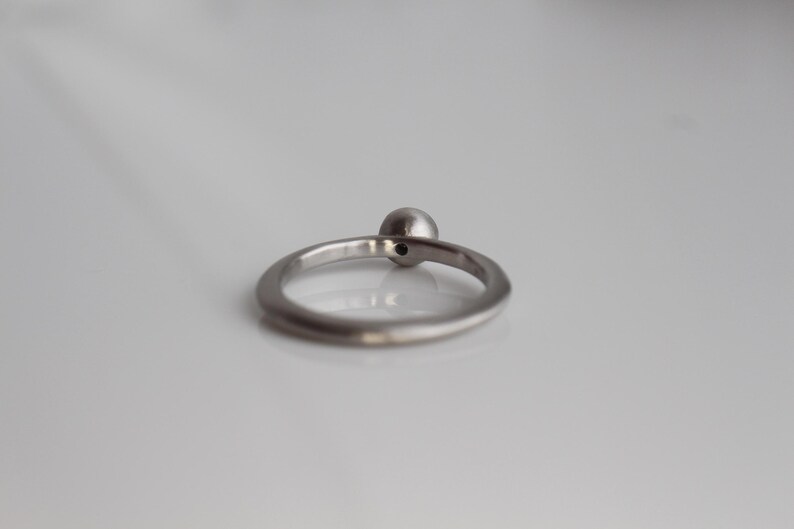 May include: A silver-toned metal ring featuring a single, small, spherical bead set at the top of the band. The ring has a smooth, polished finish and a simple, circular design that sits upright on a reflective, light-colored surface.