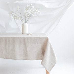 May include: A beige linen tablecloth covers a wooden table, with a white vase holding white flowers. The table is set against a white backdrop with draped fabric. The scene is bright and airy.