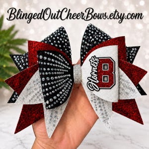 May include: A large cheer bow featuring layers of black, red, and white glitter fabric. The bow is heavily embellished with clear rhinestones and has a central rhinestone band. A white patch displays the text Hornets and a large letter B.