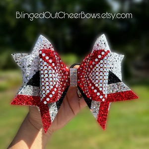 May include: A large red, white, and black cheer bow with silver glitter and rhinestones. The bow is tied with a black elastic band.