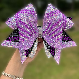 May include: A large cheer bow featuring layers of purple glitter fabric and black material, heavily embellished with iridescent and purple rhinestones. The center is wrapped in a band of silver rhinestones. Text reads BlingedOutCheerBows.etsy.com.