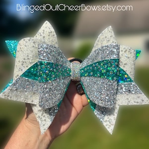 May include: A large hair bow featuring layers of white, silver, and teal glitter fabric. The center is embellished with a band of clear rhinestones. The text BlingedOutCheerBows.etsy.com is visible at the top of the image against a blurred background.