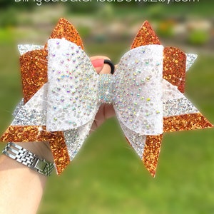 May include: A large, glittery cheer bow in shades of orange, silver, and white. The bow is embellished with sparkling rhinestones and has a black elastic band. The text "BlingedOutCheerBows.Etsy.com" is visible.