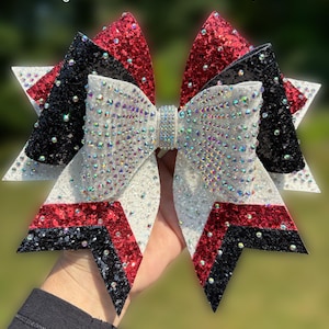 May include: A large cheer bow featuring layers of red, black, and white glitter fabric. The bow is heavily embellished with iridescent rhinestones across the surface and a central rhinestone-covered knot. Text reads BlingedOutCheerBows.etsy.com.