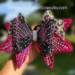 May include: A large pink, black, and gold cheer bow with rhinestones. The bow has a leopard print pattern and is tied with a black elastic band.