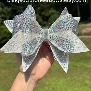 May include: A large hair bow featuring white and silver glitter fabric layers, heavily embellished with iridescent rhinestones across the surface and a central rhinestone-covered band. The text BlingedOutCheerBows.etsy.com is visible at the top.
