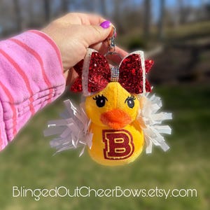 May include: A yellow plush duck keychain with a red and white glitter bow, a white pom-pom, and a maroon letter "B" on its chest. The keychain is attached to a silver ring.
