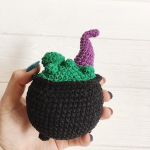 May include: A crocheted cauldron with a green top and a purple handle. The cauldron is black and has a textured surface.