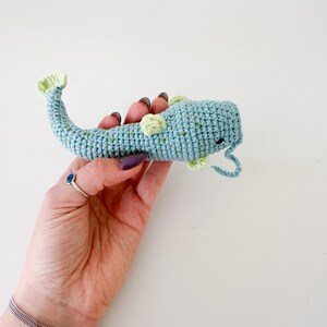 以下が含まれることがあります： 青と緑の色合いの手作りの鉤針編みの魚のおもちゃ。おもちゃは、小さな緑色のアクセントと湾曲した尾を持つテクスチャの外観をしています。魚は白い背景に対して保持され、その詳細を示しています。