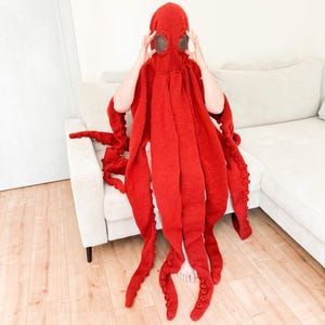 May include: A vibrant red octopus costume. The knitted hood features large, round eye openings and a series of long, tentacle-like appendages. The costume is designed to be worn, with the tentacles extending down. The overall effect is whimsical and playful.