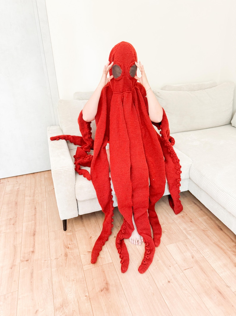 May include: A vibrant red octopus costume. The knitted hood features large, round eye openings and a series of long, tentacle-like appendages. The costume is designed to be worn, with the tentacles extending down. The overall effect is whimsical and playful.