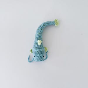 以下が含まれることがあります： 青と緑の色合いの手作りの鉤針編み魚のおもちゃ。 魚は、テクスチャパターン、小さなひれ、笑顔の顔を持つ、長く湾曲した体を持っています。 尾とひれは明るい緑色です。