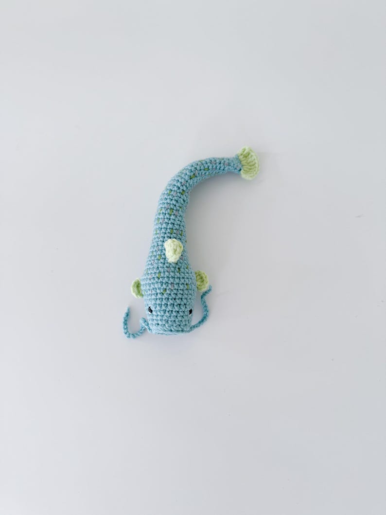 以下が含まれることがあります： 青と緑の色合いの手作りの鉤針編み魚のおもちゃ。 魚は、テクスチャパターン、小さなひれ、笑顔の顔を持つ、長く湾曲した体を持っています。 尾とひれは明るい緑色です。