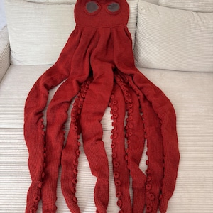May include: A knitted red octopus costume with eight long tentacles and a head with two eye holes. The tentacles have small, round details along their length. The costume is made of a soft, textured yarn.