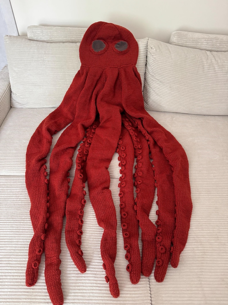 May include: A knitted red octopus costume with eight long tentacles and a head with two eye holes. The tentacles have small, round details along their length. The costume is made of a soft, textured yarn.