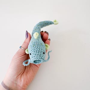 以下が含まれることがあります： 緑色のアクセントが付いた、かぎ針編みの青い魚。手作りの魚は黒い目と湾曲した尾を持っています。白い背景に、魚が手で持たれています。ユニークな手作りのアイテムです。