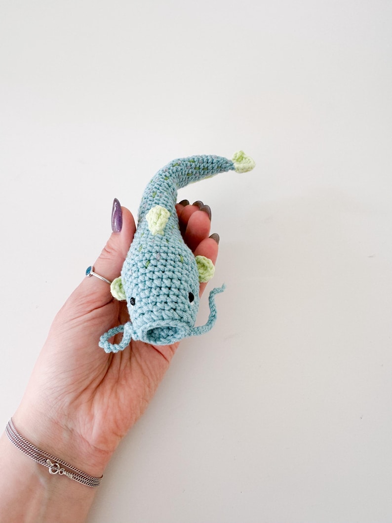 以下が含まれることがあります： 緑色のアクセントが付いた、かぎ針編みの青い魚。手作りの魚は黒い目と湾曲した尾を持っています。白い背景に、魚が手で持たれています。ユニークな手作りのアイテムです。
