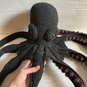 May include: A crocheted octopus plush toy with eight tentacles. The octopus is gray with purple and green accents on the tentacles. The octopus has large, red eyes.