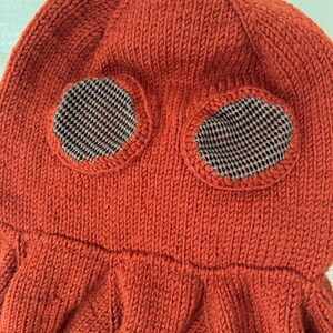 May include: A knitted orange hat with two circular eye openings. The eye openings are lined with a gray and white checkered fabric. The hat has a textured, ribbed design and a flared bottom.