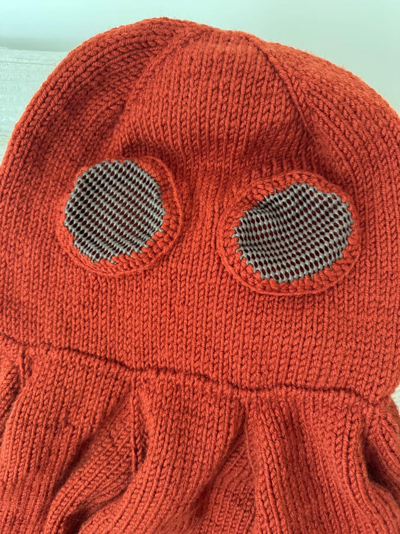 May include: A knitted orange hat with two circular eye openings. The eye openings are lined with a gray and white checkered fabric. The hat has a textured, ribbed design and a flared bottom.